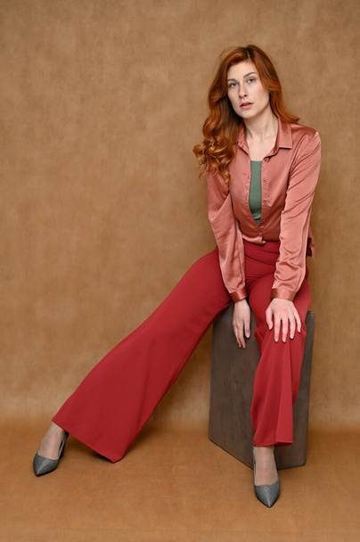 A full-length portrait of a young Caucasian red-haired woman sitting on a stand on a beige background in various poses. Nice girl with long hair dressed in red trousers and a pink blouse. - Fotó, kép