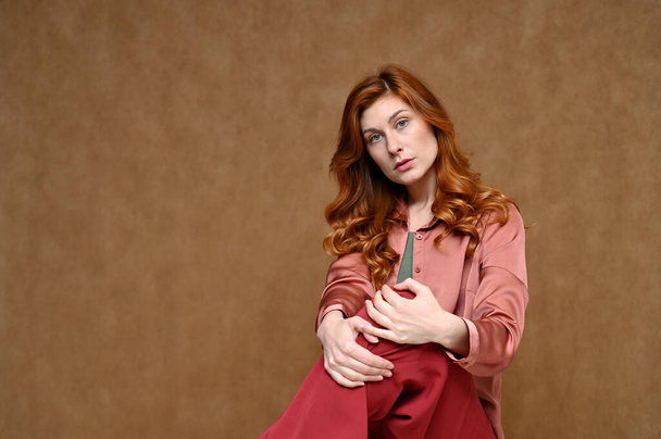 Horizontal portrait of a pretty woman with smooth emotions with long red hair posing in a pink blouse. Photo taken in the studio on a beige background. - Фото, зображення