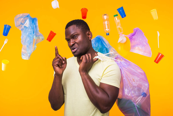 Problem of trash, plastic recycling, pollution and environmental concept - african american man carrying garbage for recycling and pointing up on yellow background. - Photo, Image