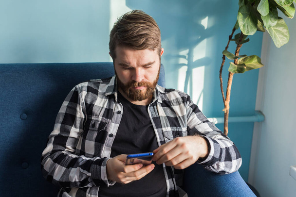 Technológiák és szabadidő koncepció - Vidám ember ül a kanapén használja okostelefonját otthon a nappaliban. - Fotó, kép