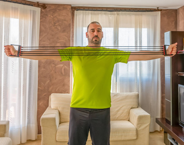 Bearded man in low shape exercising with black and green sportswear in his living room in front of the sofas on a mat - Photo, Image
