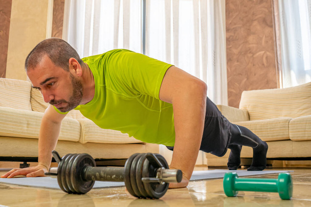Bärtiger Mann in schwacher Form beim Training in schwarz-grüner Sportbekleidung in seinem Wohnzimmer vor den Sofas auf einer Matte - Foto, Bild