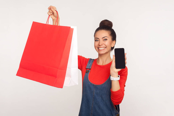 Portrait of happy shopper, stylish girl in denim overalls holding shopping bags and cell phone, advertising application for online ordering, purchase and delivery of goods. studio shot, isolated - Фото, зображення
