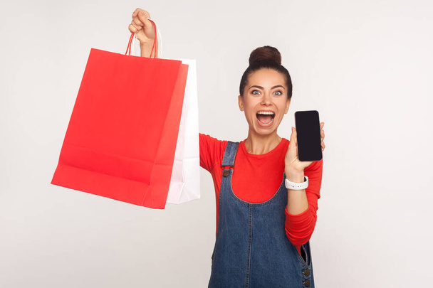 Portrait of excited amazed shopper girl in denim overalls holding shopping bags and cell phone, advertising application for online ordering, purchase and delivery of goods. studio shot, isolated - Foto, Imagen