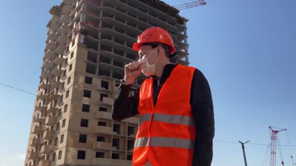 Male construction worker in overalls and medical mask coughing on background of house under construction. Concept of threat of infection. - Metraje, vídeo