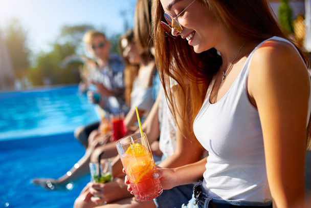 Pretty woman relax with fresh colorful cocktail glass sitting by swimming pool on sunny summer day with friends. People toast drinking beverages at luxury villa poolside party on tropical vacation. - Foto, Imagen