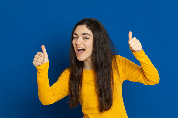 Brunette jong meisje draagt gele trui op een blauwe achtergrond - Foto, afbeelding