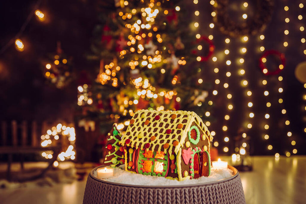 Amusante maison décorée et colorée en pain d'épice avec des fenêtres en verre caramel et une bougie illuminant l'intérieur, un arbre de Noël bokeh flou et un salon sur fond. Beaucoup d'espace de copie
. - Photo, image