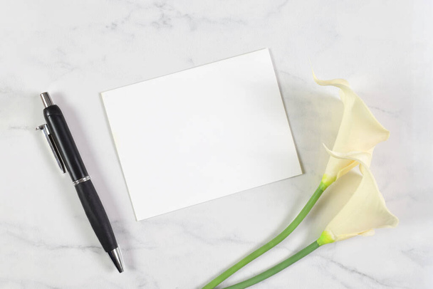 Een witte kaart rust op een witte marmeren achtergrond. Calla lelies en een sjieke pen omsingelden de blanco kaart. - Foto, afbeelding