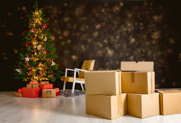Moving in to new home for Christmas concept. Focus on cardboard relocation boxes and decorated Christmas tree with presents under it on the background. Copy space on black background. - Фото, изображение