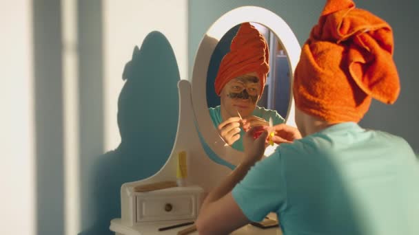 Um jovem bonito em uma máscara cosmética com uma toalha na cabeça olha para si mesmo no espelho
 - Filmagem, Vídeo