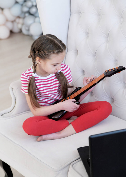  オンラインレッスン。女の子はノートパソコンを使って自宅でギターを弾くことを学ぶ。家庭教育の概念. - 写真・画像