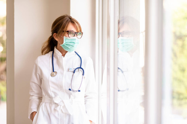 Retrato de una doctora que usa mascarilla facial para prevenir mientras está de pie junto a la ventana y mirando hacia afuera
. - Foto, Imagen