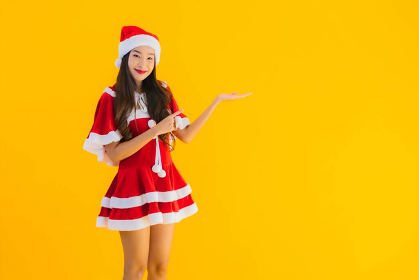 Portrét krásné mladé asijské ženy vánoční oblečení a klobouk úsměv šťastný s ostatními akcemi na žlutém izolovaném pozadí - Fotografie, Obrázek