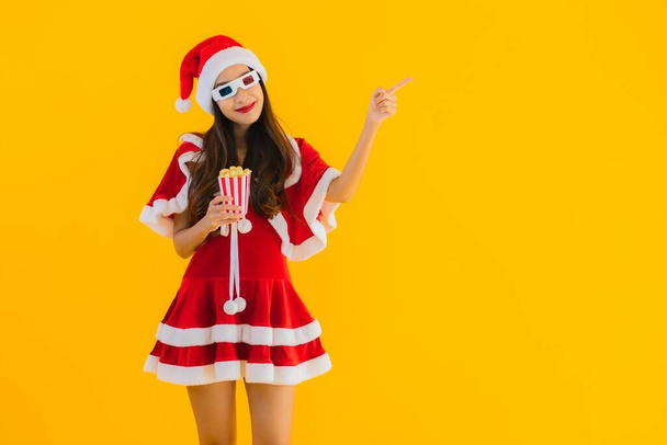 Porträt schöne junge asiatische Frau tragen Weihnachtskleidung Hut mit Popcorn und 3D-Brille bereit für Film auf gelbem Hintergrund - Foto, Bild