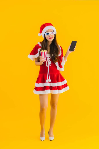 Portrait beautiful young asian woman wear christmas clothes hat with popcorn and 3d glasses ready for watch movie on yellow isolated background - Photo, Image