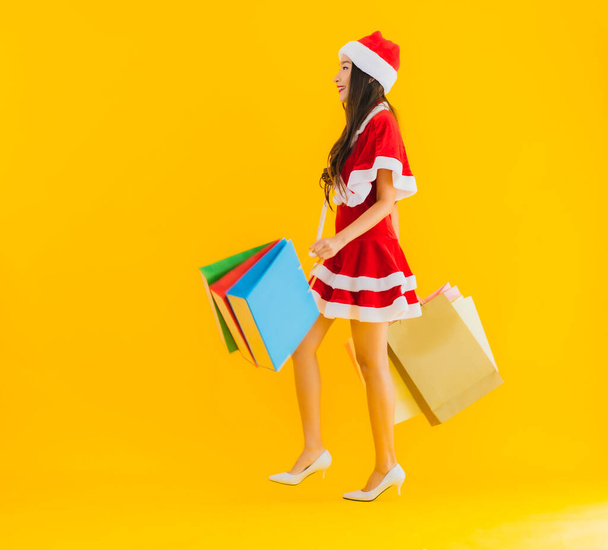 Портрет красива молода азіатка носить різдвяний капелюх з великою кількістю сумки для покупок на жовтому ізольованому фоні
 - Фото, зображення