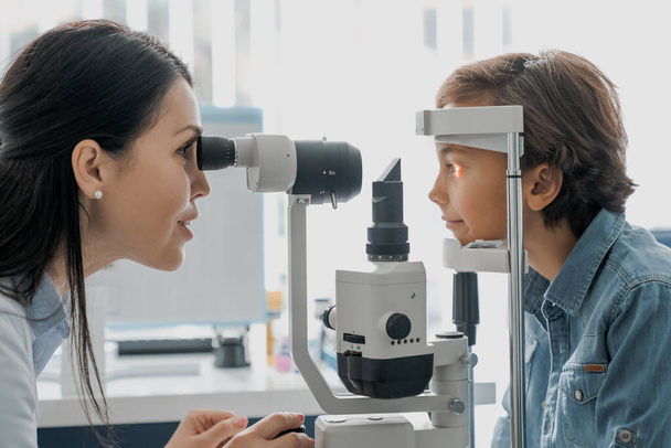 Side view shot of female doctor ophthalmologist checking the eye vision of cute child in modern clinic - Fotografie, Obrázek