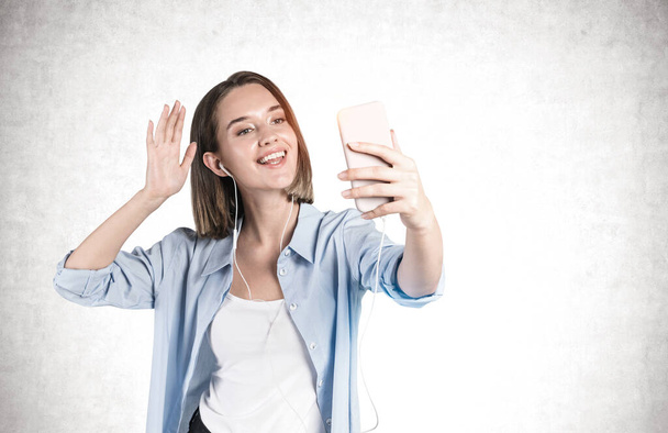 Portrait of cheerful young woman in smart casual clothes making video call or selfie and waving to the camera near concrete wall. Concept of communication and social media. Mock up - Photo, image