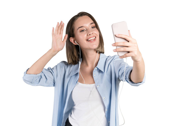 Isolated portrait of cheerful young woman in smart casual clothes making video call or selfie and waving to the camera. Concept of communication and social media - Photo, image