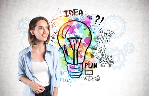 Cheerful young woman in casual clothes standing near concrete wall with colorful business idea sketch and gears drawn on it. Concept of planning and business strategy - 写真・画像