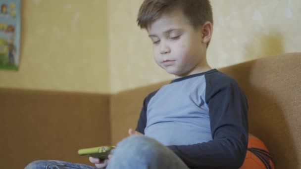 Little boy talking to parents behind camera while playing online games or educational games on smartphone sitting on the couch with ball behind him. Stay at home concept. Prores 422 - Кадры, видео