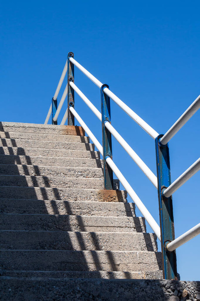 Eenvoudige betonnen trap buiten met een diagonale schaduw en een metalen reling. Heldere blauwe lucht. Heraklion, Kreta, Griekenland. - Foto, afbeelding