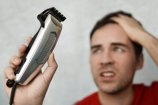 Handsome man cutting his own hair with a clipper and looks in the clipper. self-care in the conditions of global quarantine and closed hairdressers and beauty salons. electric razor - Photo, Image