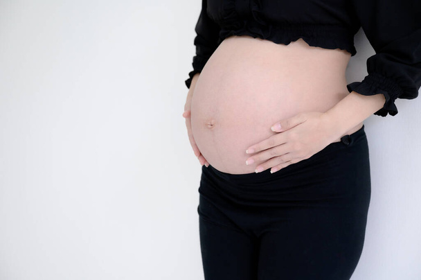 Pregnant woman in black dress and hands hold on belly on a white wall background - Foto, immagini