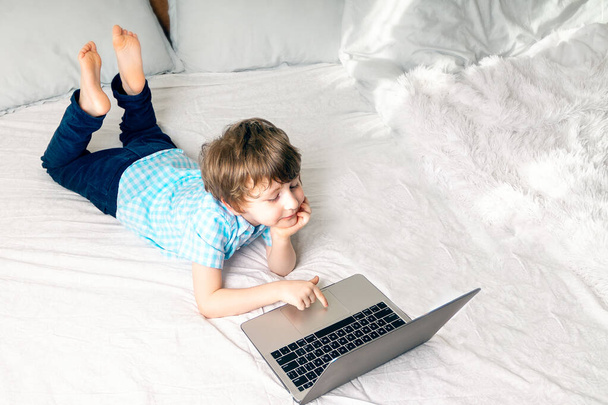 Kształcenie online na odległość. Biały szczęśliwy chłopiec z książką studiuje w domu z notatnikiem z tabletem cyfrowym i odrabia lekcje w szkole. Siedzenie na łóżku z książkami treningowymi - Zdjęcie, obraz