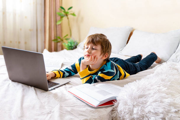 Kształcenie online na odległość. Biały szczęśliwy chłopiec z książką studiuje w domu z notatnikiem z tabletem cyfrowym i odrabia lekcje w szkole. Siedzenie na łóżku z książkami treningowymi. - Zdjęcie, obraz