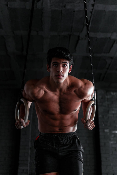 Nahaufnahme eines jungen, hemdslosen Muskelsportlers, der in einem Crossfit-Fitnessstudio Ringdips macht. Calisthenics-Profi trainiert - Foto, Bild