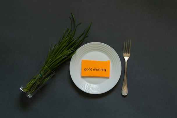 Белая тарелка куча зеленой травы и вилка на столе и следующая нота доброе утро
 - Фото, изображение