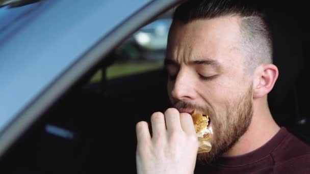 Nuori mies insdie-autossa. Muotokuva hipster kaveri pureskelee maukasta herkullista hampurilaista ja juoda paperimukista. Nauti ottaa pois aamiaisen tai lounaan autossa
. - Materiaali, video