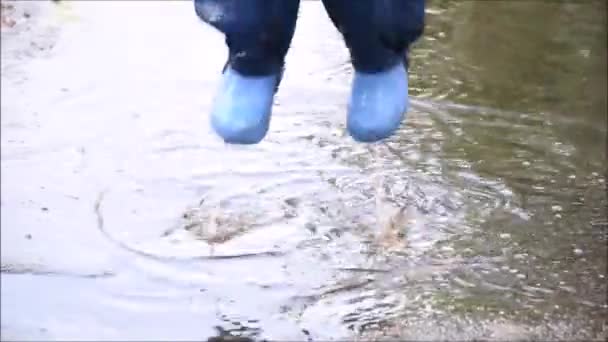 Szczęśliwe dziecko skaczące w kałuży w wodoodpornym płaszczu. Chłopiec bawi się w deszczową pogodę w jasnym płaszczu. Nogi dziecka w gumowych butach skaczą w kałuży. - Materiał filmowy, wideo