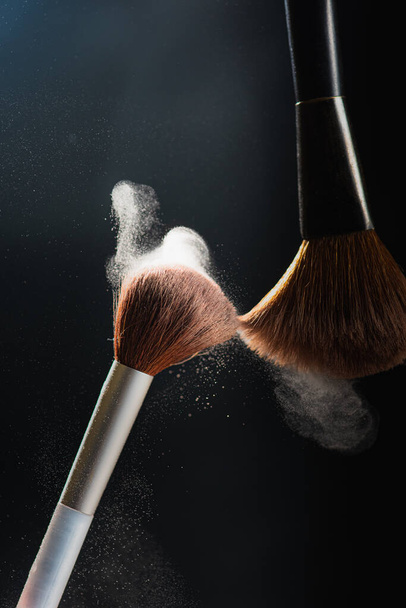 The art of visage. Horizontal closeup photo of two professional black makeup brushes with colored powder isolated on black background  - Φωτογραφία, εικόνα