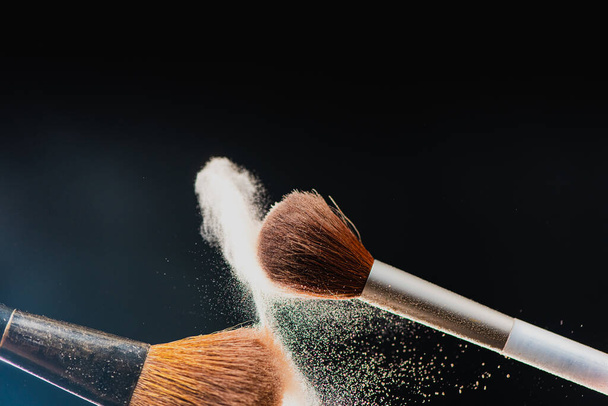 The art of visage. Horizontal closeup photo of two professional black makeup brushes with colored powder isolated on black background  - Photo, Image