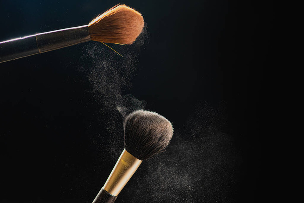 De kunst van het gelaat. Horizontale close-up foto van twee professionele zwarte make-up borstels met gekleurd poeder geïsoleerd op zwarte achtergrond  - Foto, afbeelding