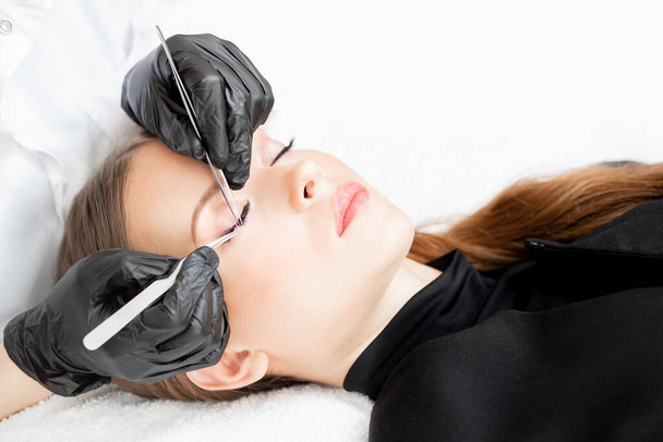 Eyelash extension procedure. Master tweezers sets fake lashes on beautiful woman - Photo, Image