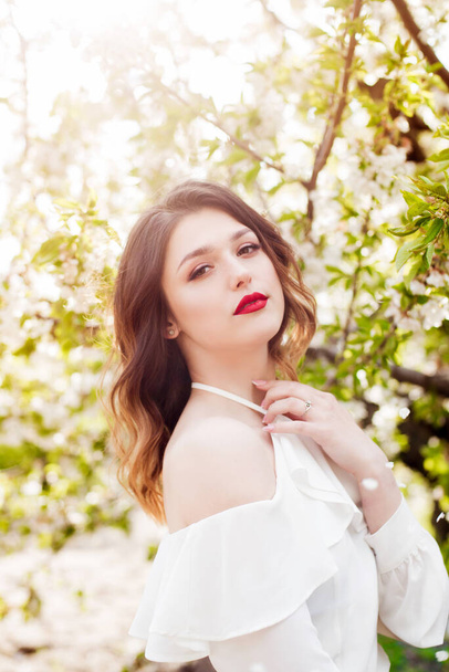 Lovely young girl in a white romantic silk blouse in spring in a blooming cherry garden in sunny weather. Spring and Fashion - Foto, Imagem