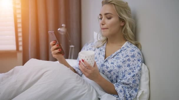 Beautiful young woman browsing smartphone and holding mug of hot beverage sitting on bed - Imágenes, Vídeo