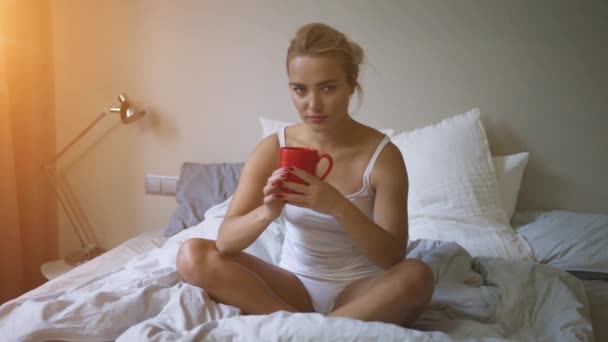 Mujer bonita en camiseta blanca y bragas sentadas en la cama y disfrutando de la bebida caliente
 - Imágenes, Vídeo