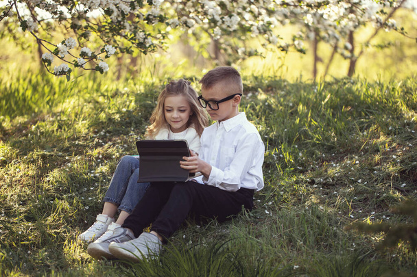 Two little Caucasian children wolk in the park with each other. Brother and sister spend time together. older brother help his younger sister with homework. Stylish clothes, cool glasses and happy faces - Fotó, kép