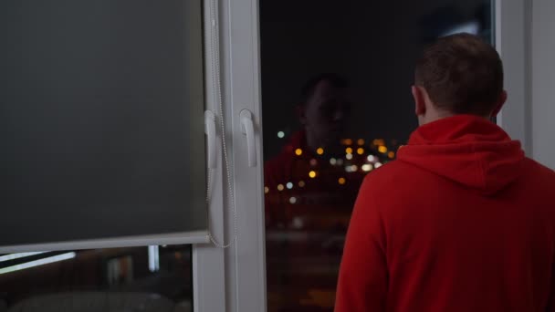 Muž v mikině stojí u okna a dívá se na noční město. - Záběry, video