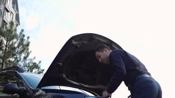Nuori mies auton ulkopuolella. Liikemies seisoo edessä avattu huppu ja pidä jakoavain korjaamiseen auton. Yrittää korjata ongelmaa yksin
. - Materiaali, video