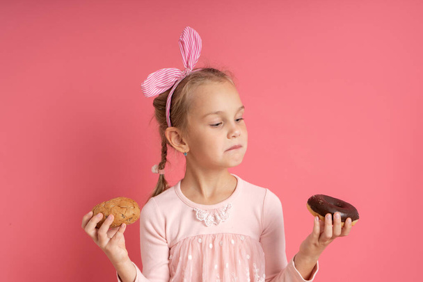 Little blonde model in dress, with headband and pigtail. Holding bun and chocolate donut in her hands. Posing on pink background - Photo, Image