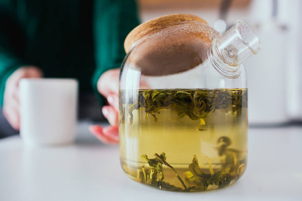 Young woman in kitchen during quarantine. Close up and cut view of teapot with green tea inside. Girl hold it and white cup in hands. Blurred background. - Fotoğraf, Görsel