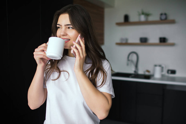 Young woman in kitchen during qiarantine. Talking on phone and drink from white cup tea or coffee. Online conversation wireless. Modern technologies and devices. - Photo, Image