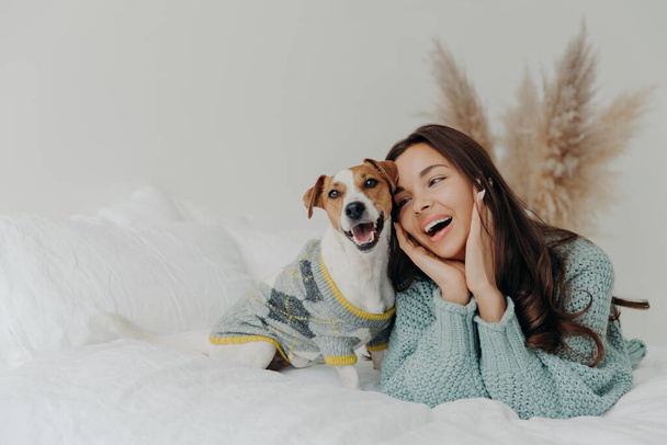 People and animals concept. Joyful pretty young woman express love to dog, spends leisure time with pet, lie together on bed, looks tenderly at animal, feels not bored with true loyal friend - Фото, изображение