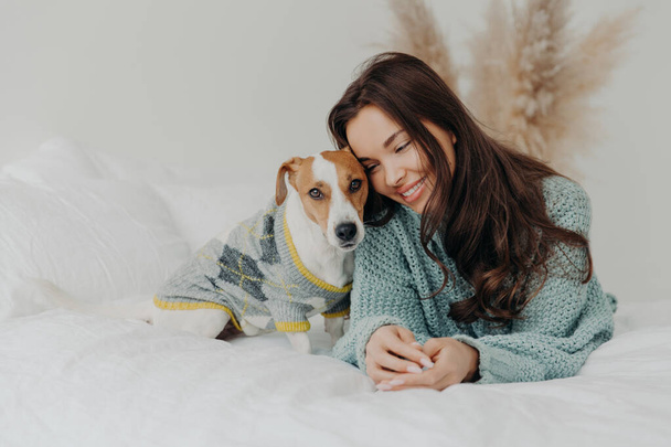 Photo of beautiful brunette woman in knitted sweater lies together wih dog on soft bed, enjoys spending time with favorite pet, cares about animals, stay at home during coronavirus quarantine - Foto, Imagen